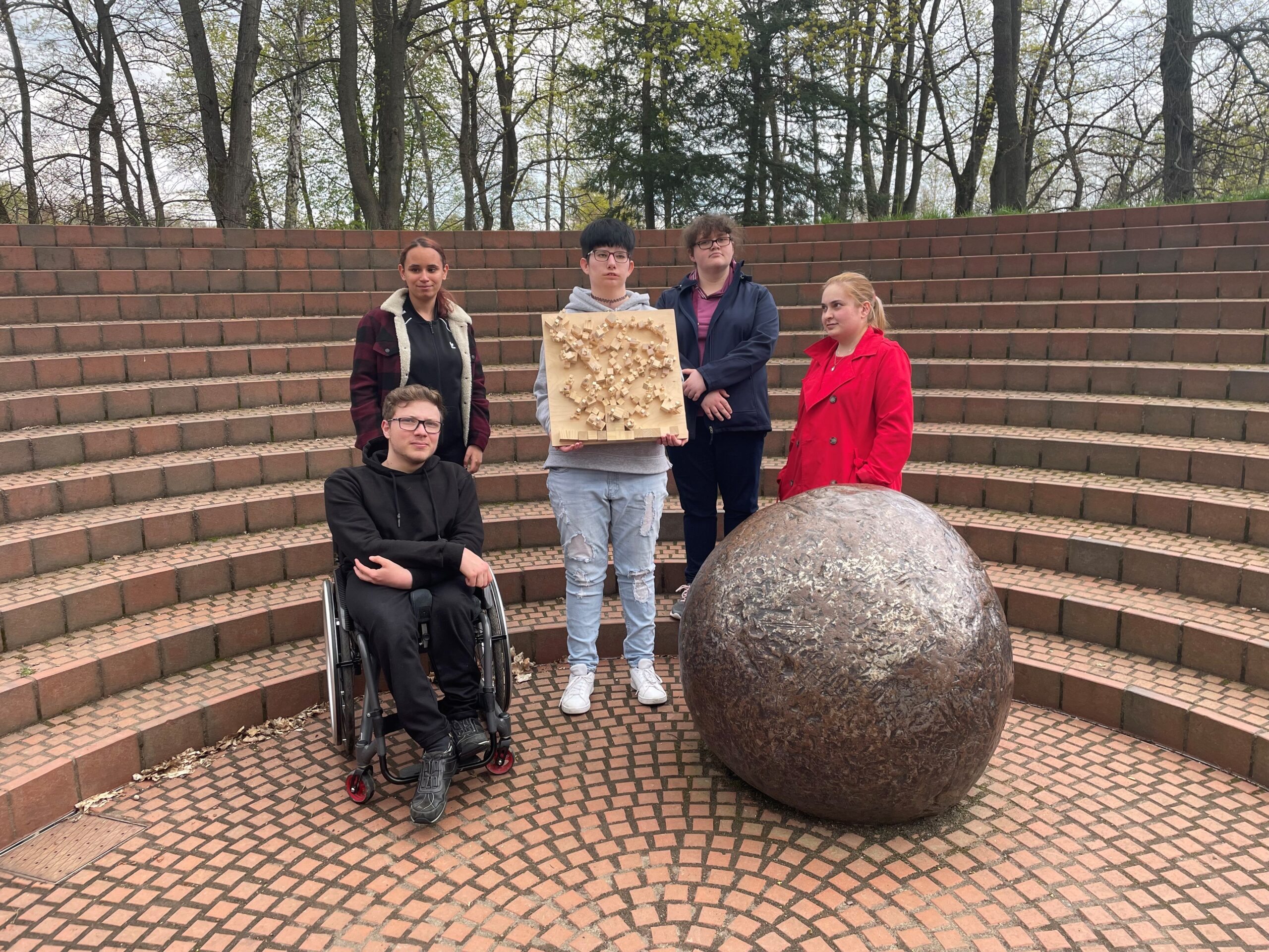Eine Gruppe von jungen BVB Teilnehmer*innen stellt im Denkmal Gedenken, ein Holzrelief vor.