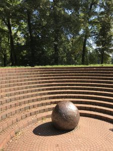 Das Denkmal GEDENKEN bei Sonnenschein. Eine deformierte Abrisskugel inmitten eines Rondells, welches an ein griechisches Theater erinnert. Im Hintergrund sind Bäume zu sehen.