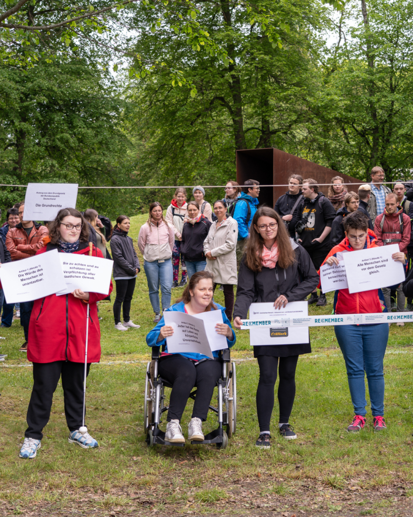 Im Vordergrund stehen Menschen mit Behinderung. Sie zeigen Artikel aus dem Grundgesetz und ein Absperrband, mit dem Tatorte der Nationalsozialisten markiert werden. Im Hintergrund sind das Denkmal GEDENKEN sowie viele Mitarbeitende und Auszubildende des SFZ zu sehen.