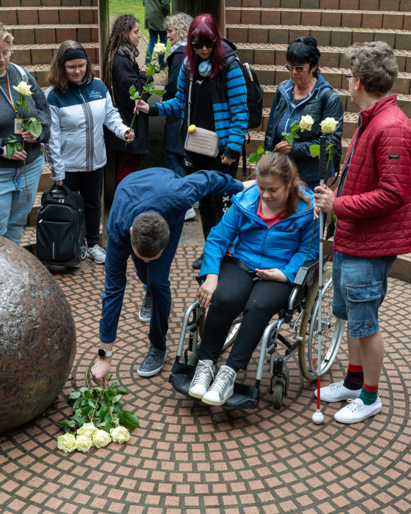 Im Denkmal GEDENKEN haben sich Menschen mit und ohne Behinderung versammelt, um weiße Rosen niederzulegen.