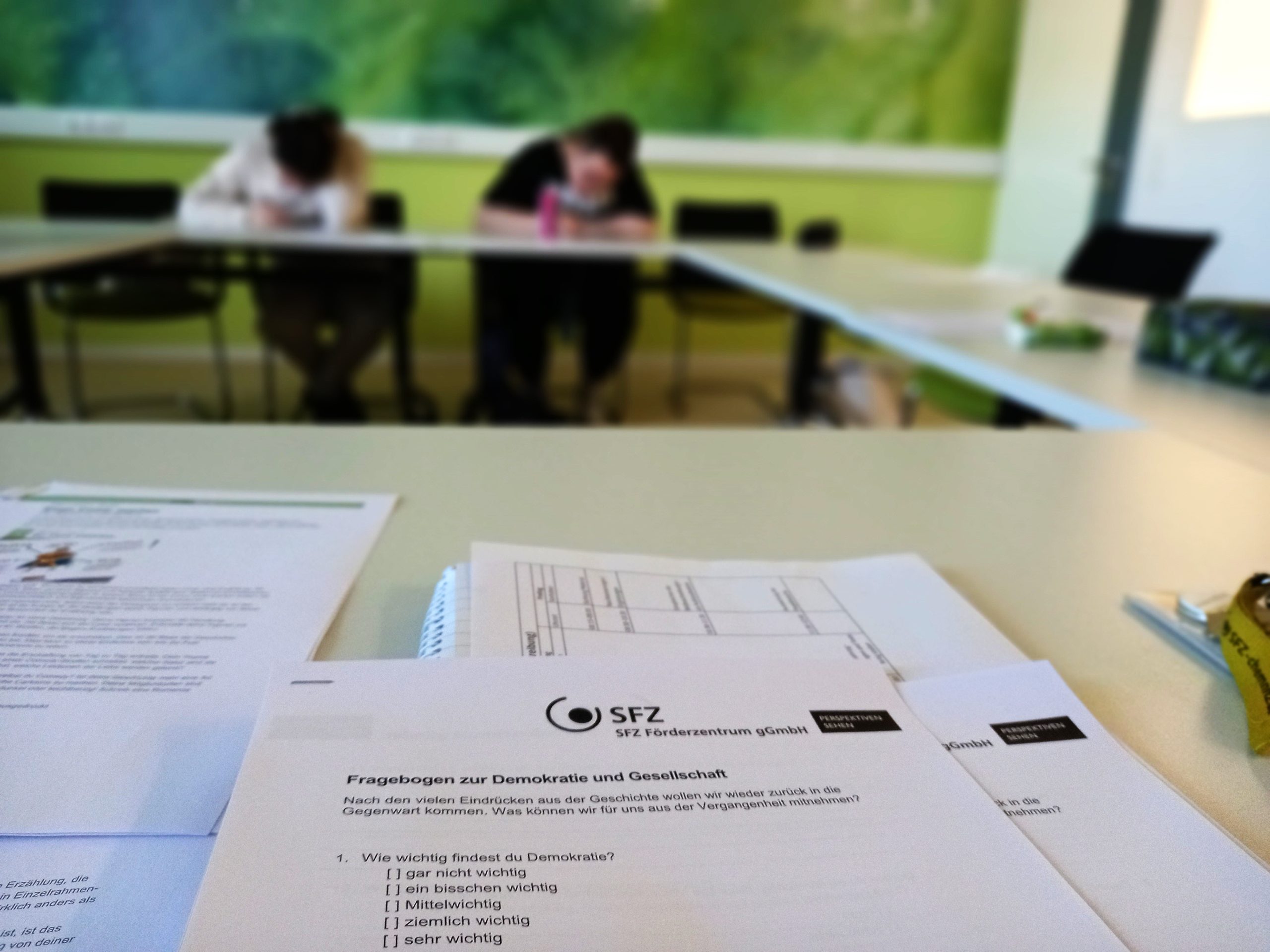 Im Vordergrund ist ein Fragebogen zu sehen. Dahinter öffnet sich der Blick in den Seminarraum, in dem junge Menschen arbeiten.