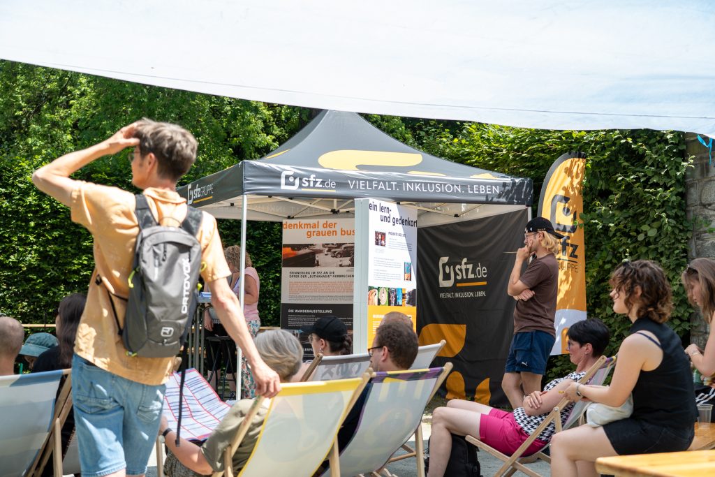 Blick aus den reihen der Liegestühle auf den Informationsstand von Unantastbar Mensch. Ein Besucher liest sich interessiert einen Aufsteller durch.