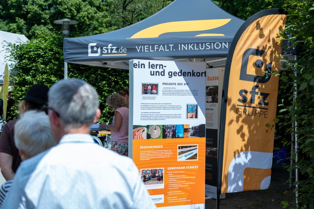 Der Informationsstand von Unantastbar Mensch mit Aufstellern, die über das Projekt informieren. Im Vordergrund sind einige Besuchende des Festivals zu sehen.