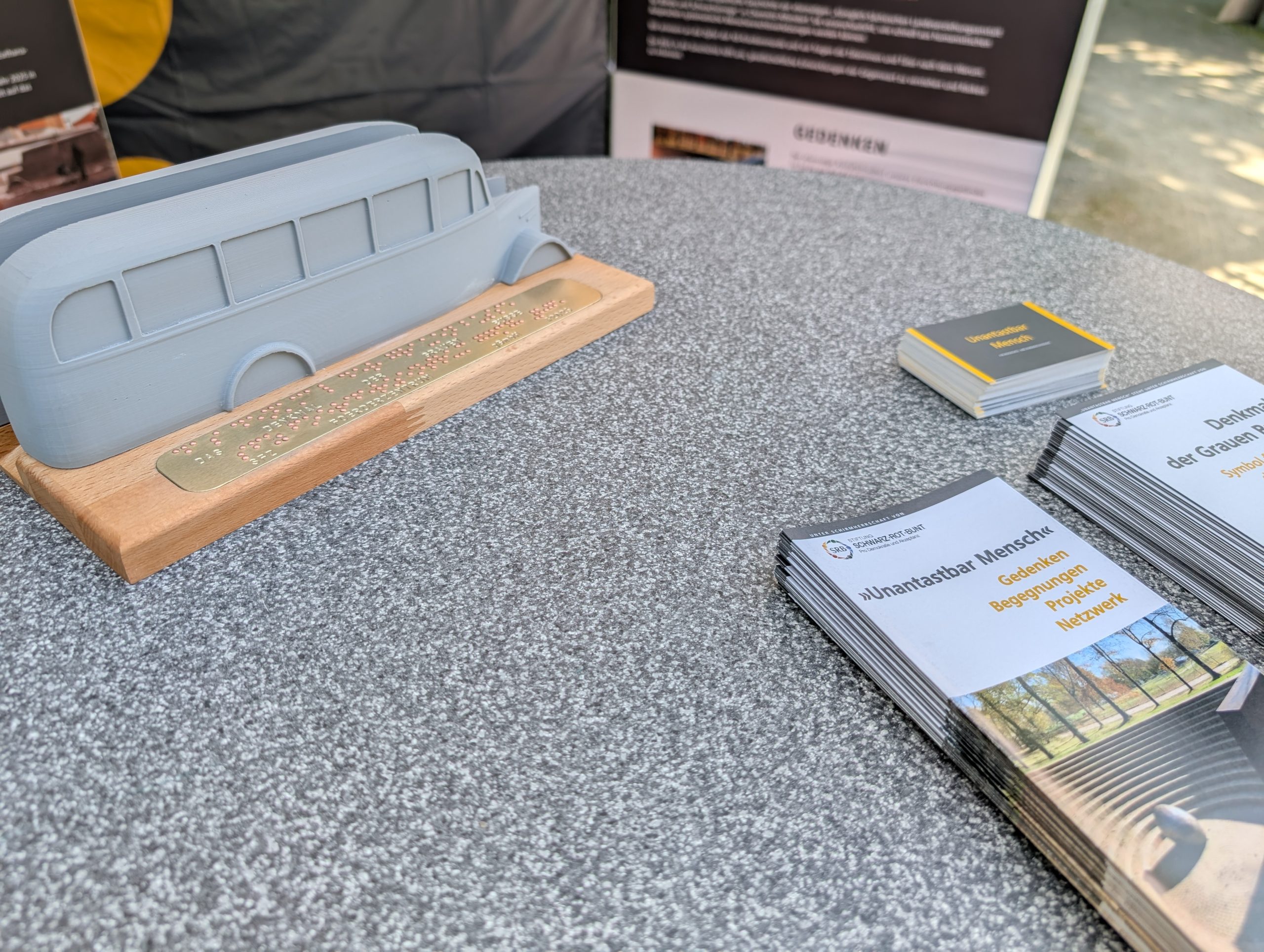 PXL_20250614_101528803 Das Modell des Denkmals der Grauen Busse mit Beschriftung in Brailleschrift und Schwarzschrift. Zu sehen sind außerdem Flyer von Unantastbar Mensch.