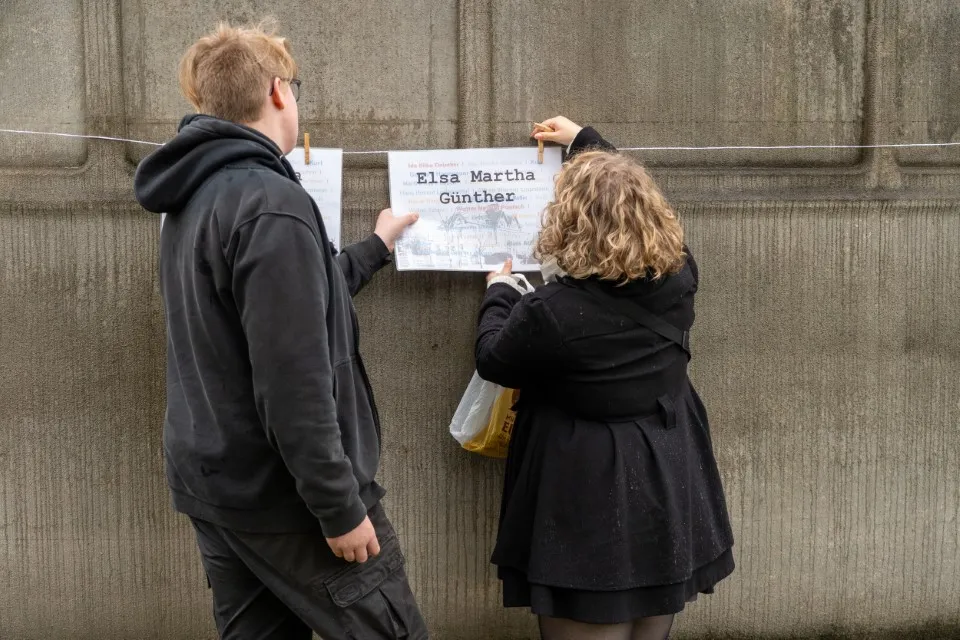 Ein Auszubildender hängt gemeinsam mit einer Mitarbeiterin ein Namensplakat am Denkmal der Grauen Busse auf.